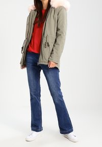 Žena v olivově zelené parce s růžovou kožušinovou kapucí, červeném tričku, modrých džínách a bílých teniskách, stojící na čistě bílém pozadí.