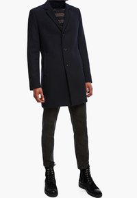 Mørkeblå uldfrakke med skræddersyet design, enkeltbrystet lukning og to sidelommer. Båret over en struktureret striktrøje og mørke bukser.