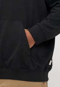 Male hand resting inside a black hoodie’s front pocket, beige pants visible below, showing casual clothing details and textures.