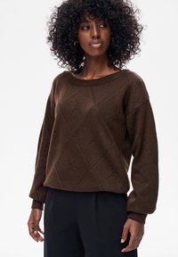 Kobieta z kręconymi włosami, ubrana w brązowy, wzorzysty sweter z dzianiny i czarne spodnie, patrząca lekko na bok na białym tle.