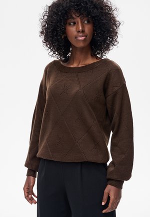 Kobieta z kręconymi włosami, ubrana w brązowy, wzorzysty sweter z dzianiny i czarne spodnie, patrząca lekko na bok na białym tle.