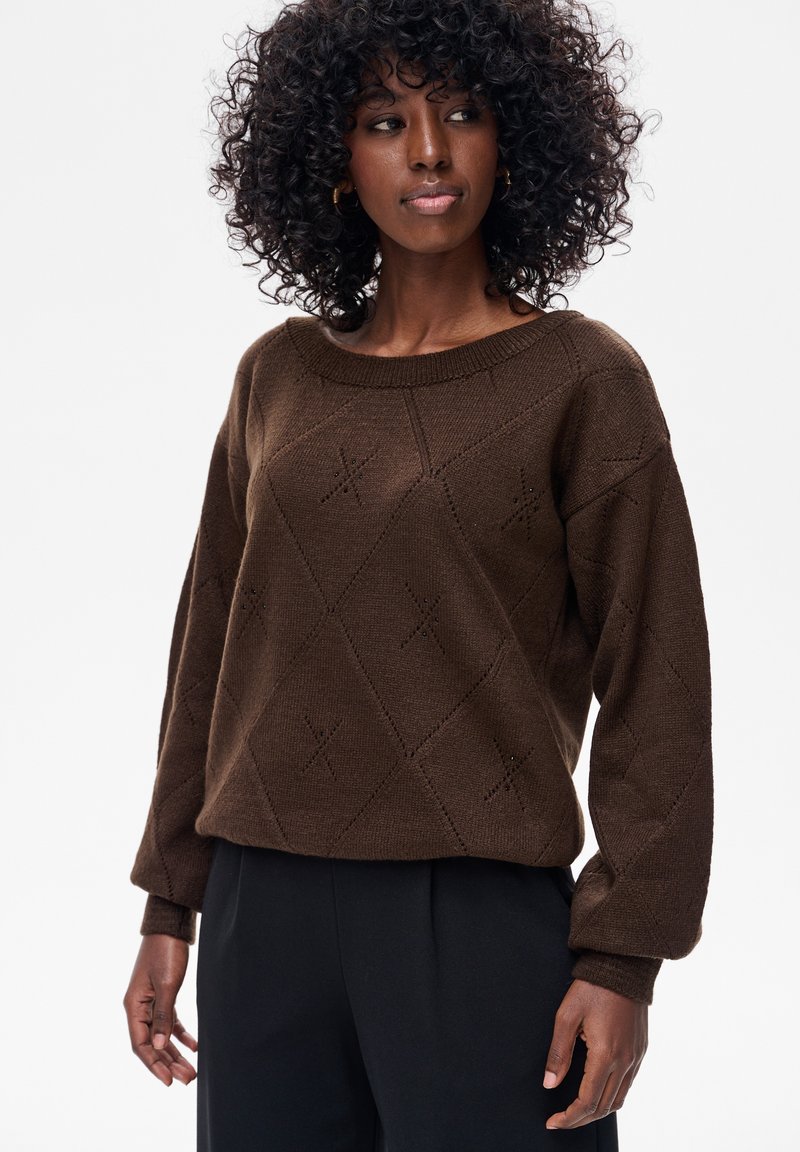 Kobieta z kręconymi włosami, ubrana w brązowy, wzorzysty sweter z dzianiny i czarne spodnie, patrząca lekko na bok na białym tle.