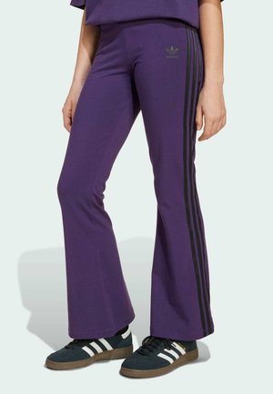 Lila ausgestellte Hosen aus weichem Material mit schwarzen Seitstreifen und dezentem Adidas-Logo. Kombiniert mit schwarzen Sneakers mit weißen Streifen.