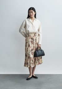 Cremefarbene Bluse zum Knöpfen mit plissiertem Blumensrock in Beige, schwarze Ballerinas und eine kleine schwarze Handtasche mit runden Griffen und minimaler Hardware.