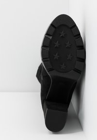 Semelle d'une chaussure noire avec six motifs de crampons en forme d'étoile, dotée d'un talon bas et épais, posée verticalement contre un angle blanc.