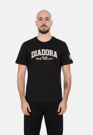 Uomo calvo con barba che indossa una maglietta nera Diadora "1948 Run Valley" e pantaloni neri, in piedi davanti a uno sfondo bianco semplice.