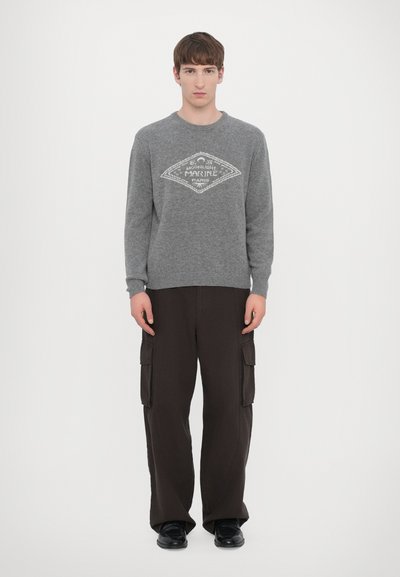 Pullover grigio in lana con una grafica bianca a forma di diamante, abbinato a pantaloni cargo neri larghi con tasche laterali.