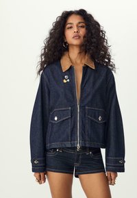 Veste en denim avec un col en velours côtelé marron, fermeture éclair à l'avant, deux poches poitrine et des épinglettes décoratives. Le design présente un tissu bleu foncé et des coutures visibles.