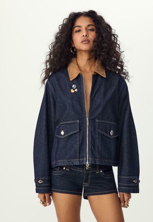 Denimtakki, jossa on ruskea vakosamettikaulus, etuvetoketju, kaksi rintataskua ja koristepinssejä. Suunnittelu sisältää tummansiniset kankaat ja näkyvät saumat.