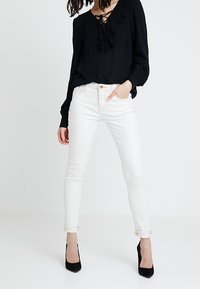 Jeans skinny bianchi con vita media, risvolti alle caviglie, abbinati a una blusa nera con collo arricciato e dettagli con lacci. Tacchi neri.