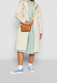 Trench beige con grandi bottoni, abbinato a un vestito a righe verde e bianco. Strati di sneakers blu e calze bianche completano il look.