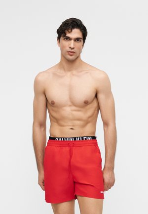 Inges, rövid, göndör hajú férfi piros fürdőnadrágban és fekete Calvin Klein alsónadrág derékrésszel, egyszerű háttér előtt.