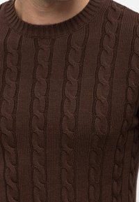 Maglione a maglia marrone con collo a girocollo a coste e motivi a treccia in rilievo. La texture è spessa e strutturata, offrendo calore e comfort.