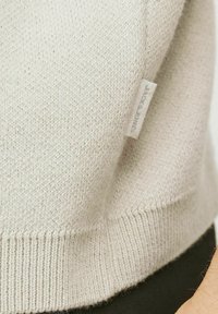 Beige stickad tröja med ribbad nederkant och en liten tygetikett som visar svag "JACK & JONES"-logotyp.
