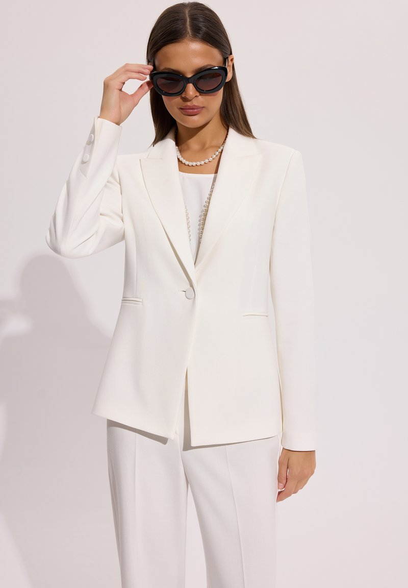 Kobieta w białej marynarce i spodniach, poprawiająca czarne okulary przeciwsłoneczne, z perłowym naszyjnikiem na jednolitym tle.