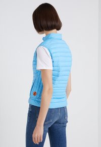 Gilet matelassé bleu clair avec un col haut, un design avec des coutures horizontales et un logo orange circulaire sur le côté gauche. Porté par-dessus une chemise blanche.