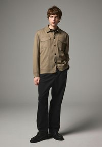 Long-sleeve khaki shirt with two chest pockets, black buttons, and a straight cut, paired with black trousers and black shoes.