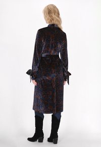 Fluwelen wikkeljas met een blauw en roestkleurig paisleypatroon, lange mouwen met gestrikte manchetten en een ceintuur in de taille. Vlakke kraag, halverwege de kuit.