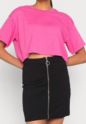 Kobieta w jasnoróżowym krótkim T-shircie i czarnej spódnicy z wysokim stanem, z zamkiem błyskawicznym z przodu i ozdobnym pierścieniem.