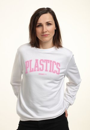Femme aux cheveux bruns mi-longs portant un sweat-shirt blanc avec un texte rose "PLASTICS Mean Girls", debout les mains dans le dos.
