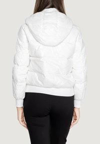 Giacca puffa bianca con cappuccio, caratterizzata da un design trapuntato, polsini e orlo a costine e una finitura lucida. Indossata sopra pantaloni neri.