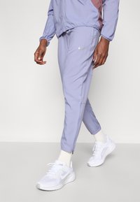 Άνδρας περπατάει με ανοιχτό μωβ φόρμα Νίκε, ταιριαστό μπουφάν, λευκές κάλτσες και λευκά αθλητικά παπούτσια Νίκε σε ουδέτερο φόντο.