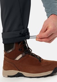 Brun vandringskänga med mesh- och mockamaterial, som har svarta snören och stöd för vristen; hand som justerar svart elastik vid byxslutet.