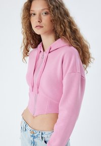 Sudadera rosa corta con cremallera en el frente, textura suave, mangas largas y un bolsillo tipo canguro. Combinada con unos jeans de denim azul claro.