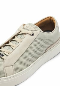 Sneaker beige chiaro con superiore in mesh e suede, corpo testurizzato, lacci piatti e una suola bianca spessa con dettagli accentuati discreti.