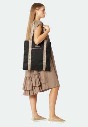Sac fourre-tout noir avec une poche zippée à l'avant et des straps textured beiges arborant un branding en relief. Modèle portant une robe superposée de couleur neutre.