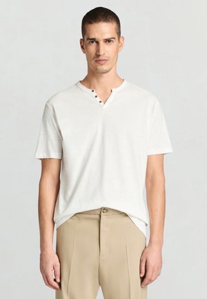 Uomo con capelli corti che indossa una maglietta henley bianca a maniche corte e pantaloni beige, in piedi contro uno sfondo chiaro e uniforme.