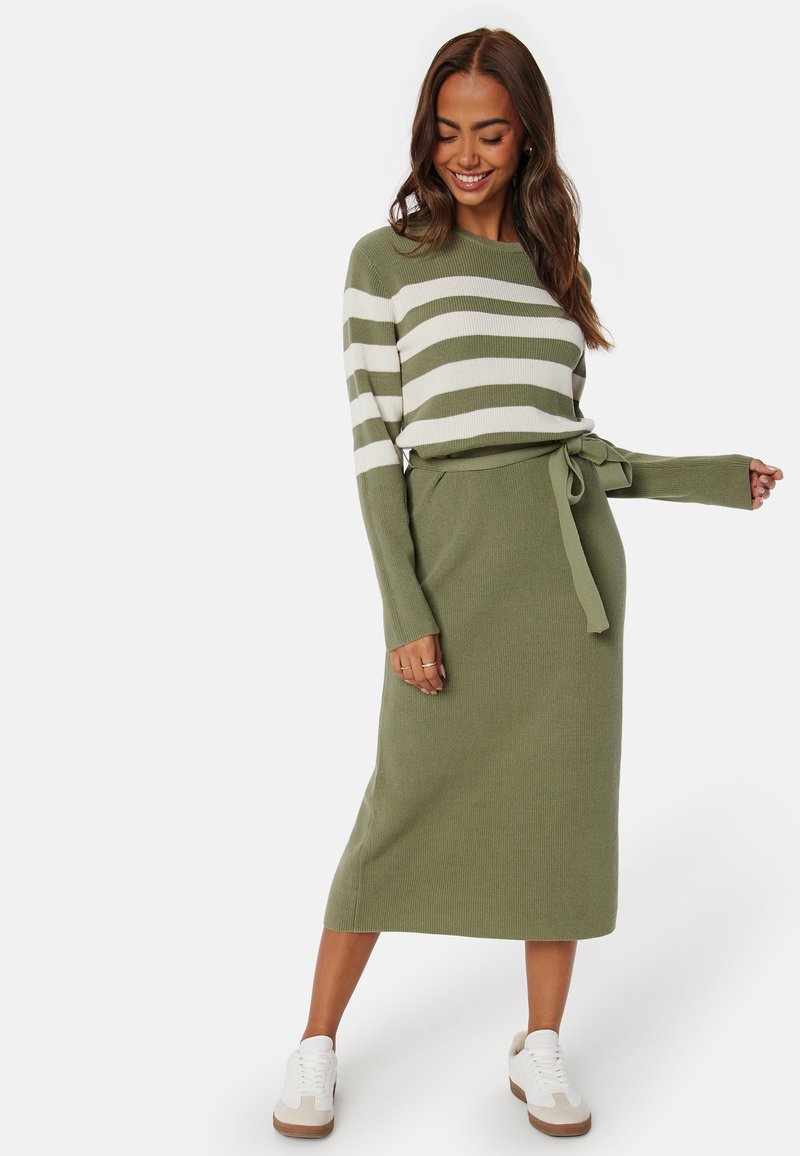 Abito lavorato a maglia verde oliva con strisce bianche, dotato di una vita con cintura e maniche lunghe, abbinato a sneaker bianche per un look casual.