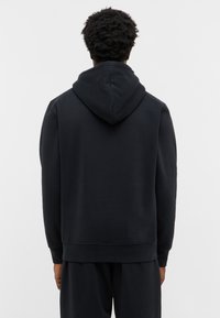 Mężczyzna z krótkimi kręconymi włosami, ubrany w prostą czarną bluzę z kapturem i czarne spodnie, pokazany z tyłu na jednolitym tle.