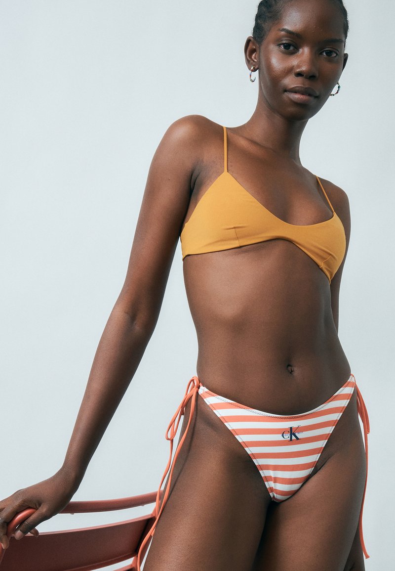 Naine, kes kannab sinepikollast bikiinikõrvalt ja triibulisi oranži-valgeid bikiinipõhjaosasid, seisab punase tooli kõrval heleda tausta ees.