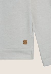 Maglietta a maniche lunghe grigia chiara con una texture morbida, caratterizzata da un piccolo logo quadrato in bronzo sul bordo e un dettaglio con spacco laterale.