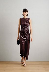 Αμάνικη, εφαρμοστή, μπορντώ φόρεμα με ραφές κατά μήκος του μπροστινού μέρους, τονίζοντας μια πλάγια σχισμή, σε συνδυασμό με μαύρα αξεσουάρ και σανδάλια.