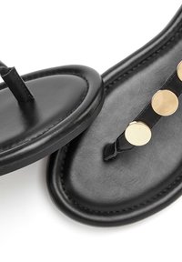 Črne usnjene sandale z ravnim podplatom, obliko, ki se prilega prstom, in tremi kovinskimi zlatimi okroglimi dodatki ob trakovi. Gladka tekstura.