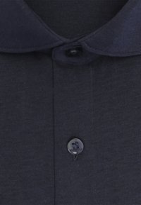 Chemise bleu foncé avec un col arrondi fermé par un bouton du haut et un bouton noir supplémentaire visible en dessous sur le tissu.