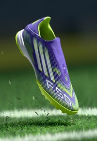Purple and neon green soccer boot with textured surface, aerodynamic design, prominent three stripes, and conical studs.