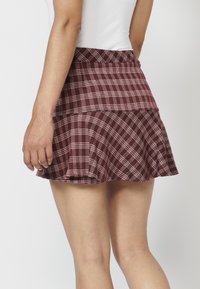 Femme portant une jupe plissée à carreaux bordeaux et un haut blanc sans manches, vue de dos sur un fond uni.