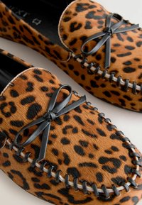 Mocassini con stampa leopardata realizzati in pelliccia sintetica marrone texture, con fiocchi in pelle nera e cuciture decorative lungo i bordi.