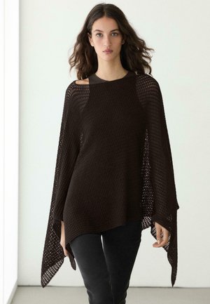 Donna con lunghi capelli castani, indossa un poncho lavorato a maglia marrone scuro e ampio sopra un top nero e pantaloni neri, camminando in una stanza chiara.