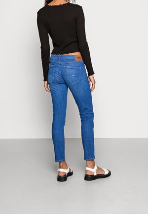 Blå denim skinny jeans med et brunt lædermærke på den bageste talje, parret med en sort langærmet top og hvide sandaler.