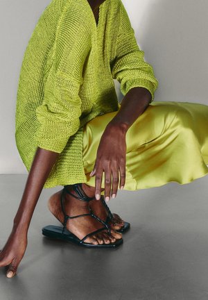 Stickad grön tröja med lös passform och ribbade muddar, kombinerad med en glänsande gul satinkjol. Svarta remsandaler på bara fötter.
