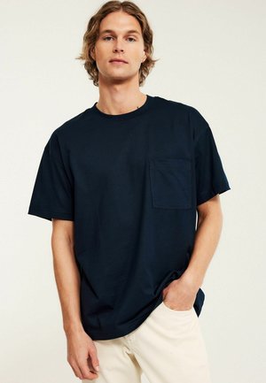 Junger Mann mit hellbraunen Haaren steht vor einem einfarbigen Hintergrund und trägt ein schwarzes T-Shirt mit kurzen Ärmeln und Tasche sowie helle beige Hosen.