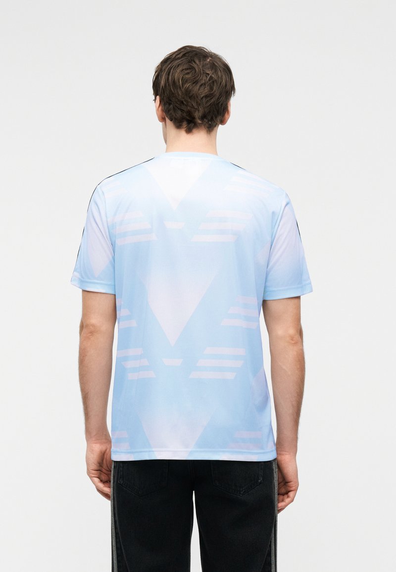 Mann trägt hellblaues Hemd mit dezenten geometrischen Mustern, zeigt den Rücken, kombiniert mit schwarzer Hose vor einfarbigem Hintergrund.