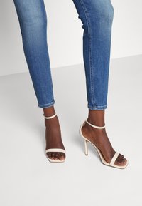 Beige högklackade sandaler med fyrkantig tå, smal ankelrem och slät läderstruktur, kombinerat med slim fit blå jeans.