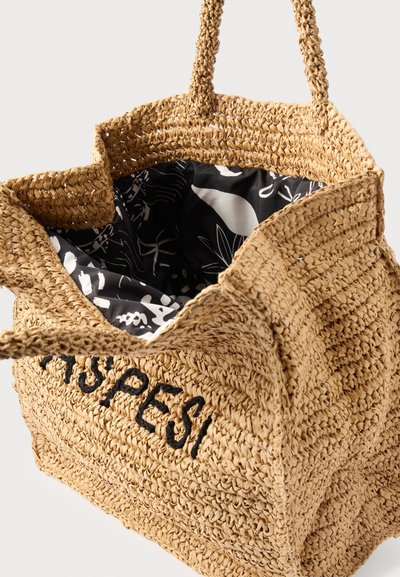 Πλεκτή τσάντα ώμου από ψάθα με μαύρη κεντημένη επωνυμία, πλεξούδες χειρολαβές και ύφασμα επένδυσης με μαύρο-άσπρο μοτίβο στο εσωτερικό.