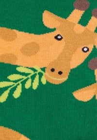 Strickstoff mit einem Giraffenmuster in Beige und braunen Akzenten, vor einem grünen Hintergrund, der grüne Blätter hält.