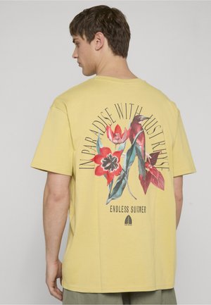 Mand iført gul t-shirt med røde blomster, en fugl og teksten "In Paradise With Just Ruise" og "Endless Summer" på bagsiden.